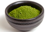 Fresh matcha green tea powder in traditional black ceramic bowl on white background, close-up view showing vibrant color and fine texture of premium Japanese tea.