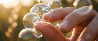 Hands gently touching a soap bubble in warm golden sunlight, with multiple iridescent bubbles floating in a soft bokeh background.