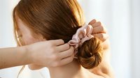 Woman with reddish-brown hair styling her hair into a bun with a scrunchie, demonstrating hairstyling techniques for beauty and personal grooming tutorials.