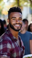 Young African American man with bright genuine smile wearing plaid shirt outdoors on sunny day, natural bokeh background creates warm welcoming atmosphere.