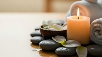 Spa wellness scene with burning candle, hot stones, towels and flower petals creating peaceful relaxation atmosphere for massage therapy treatments.