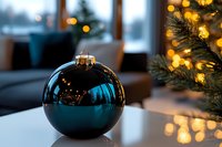 Elegant blue Christmas ornament ball with golden cap on white surface near decorated tree with warm lights in cozy modern living room interior.