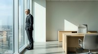 Middle-aged man in dark business suit standing by floor-to-ceiling windows in modern high-rise office, gazing at cityscape view. Corporate professional contemplating career decisions.