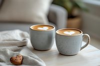 Two ceramic cups with heart-shaped latte art on coffee drinks, placed on white linen cloth with chocolate cookie, creating cozy morning atmosphere.
