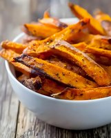 Roasted sweet potato wedges seasoned with herbs and spices served in white ceramic bowl on rustic wooden table, close-up view of healthy vegetable side dish.