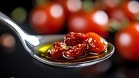 Roasted cherry tomatoes in olive oil on silver spoon with blurred fresh tomatoes background, macro food photography showing texture and shine.