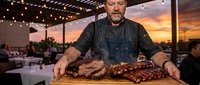Middle-aged Caucasian chef in denim apron presenting grilled meat platter on wooden board at sunset rooftop restaurant terrace with string lights overhead.
