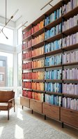 Modern home library with color-coordinated books on wooden shelves, leather chair and large windows creating bright reading space for interior design.