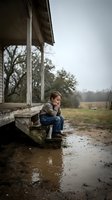 Young boy sitting on wooden porch steps during rainy day, contemplating outdoors in rural countryside setting with muddy puddles and bare trees.