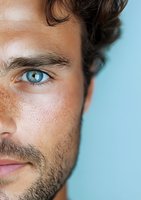 Close-up portrait of young Caucasian male with intense blue eyes, dark curly hair, and natural freckles against light blue background, showing half of face in detail.