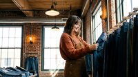 Woman with dark hair and medium complexion browsing denim jeans on rack in modern retail clothing store with brick walls, pendant lighting, and natural window light for fashion retail and shopping concepts.