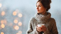 Young Caucasian woman in cozy knitted sweater and scarf holding cup of hot drink while looking away against blurred winter lights background.