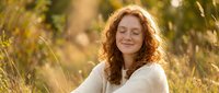 Peaceful redhead woman with curly hair meditating outdoors in golden sunlight, eyes closed in serene moment of mindfulness and relaxation in natural setting.