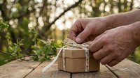 Hands tying twine around kraft paper gift box on wooden table with blurred green foliage background, suitable for holiday, birthday, and eco-friendly packaging concepts.