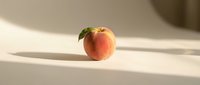 Fresh ripe peach with green leaf on white surface in soft natural sunlight creating elegant shadows for healthy eating and organic fruit concepts.