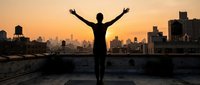 Person celebrating success with raised arms on urban rooftop at golden hour sunset with city skyline backdrop for motivational content.