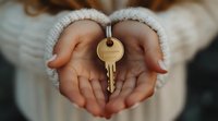 Female hands in warm white sweater holding golden house key, symbolizing home ownership, real estate purchase, or new beginnings. Soft focus background creates intimate mood.