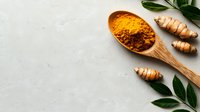Fresh turmeric powder in wooden spoon with raw turmeric roots and green curry leaves on light gray concrete background, copy space for text.