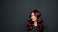 Young woman with long burgundy wavy hair and fair complexion against dark gray background, suitable for beauty, haircare, and salon marketing materials.