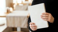 Woman holding blank white book or notebook in cozy kitchen setting with warm natural lighting for mockup design projects.