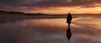 Silhouetted surfer holding board standing in shallow ocean water at sunset with dramatic golden and purple sky reflecting on wet sand beach.