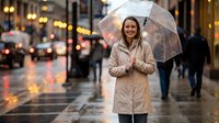 Young woman with long brown hair and fair complexion wearing a beige coat holds a transparent umbrella on a rainy urban street with blurred pedestrians and warm city lights in background.