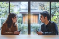 Young Asian couple sitting at cafe table with smartphones, sharing casual conversation by window with natural daylight and garden view background.