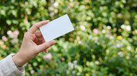 Hand holding blank white business card against blooming garden with green foliage and pink flowers. Mockup for branding, marketing, and corporate identity design projects.