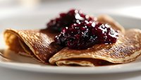 Homemade thin crepes served on white plate with sweet berry jam topping, close up view showing delicate texture and golden brown color of traditional breakfast.