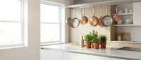Modern white kitchen interior with copper pots hanging from rail, fresh herbs in terracotta planters on marble countertop, natural sunlight streaming through window.