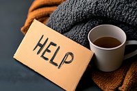 Cardboard sign with help message next to coffee cup and warm knitted scarf on dark background, concept of poverty and homelessness assistance.