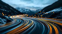 Long exposure night shot of winding mountain highway with light trails from vehicles creating glowing curves against snowy winter landscape and city lights.