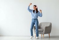 Happy young Asian woman celebrating success with raised arms wearing casual denim outfit standing near modern gray armchair in bright living room.