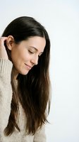 Young woman with long dark hair and fair complexion wearing beige knit sweater, looking down with serene expression, hand near ear, isolated on white background for portrait and wellness use.