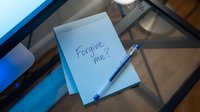 Handwritten forgive me note on blue sticky pad with pen on glass desk, expressing apology, regret, and emotional vulnerability in relationships.
