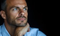 Confident middle-aged Caucasian man with blue eyes and beard wearing light blue shirt gazes thoughtfully against dark background, expressing contemplation and success.