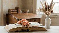 Cozy reading corner with open book, pink roses and pampas grass in vase on wooden desk near window creating peaceful home study atmosphere.