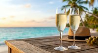 Two champagne flutes filled with sparkling wine on wooden table at tropical beach resort during golden hour sunset with palm trees and turquoise ocean view.