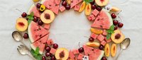 Colorful summer fruit wreath with watermelon slices, peaches, and cherries arranged in a circle on a light linen background.
