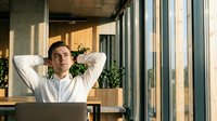 Young Caucasian businessman relaxing at office desk with laptop, hands behind head, enjoying a break in modern sunlit workspace.