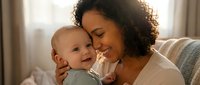 Happy mixed-race mother holding smiling baby boy in cozy home setting. Tender maternal bonding moment with infant son. Family love and parenting concept.