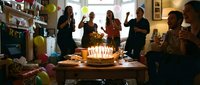 Friends celebrating a birthday party indoors with a candle-lit cake, colorful balloons, and festive decorations in a cozy living room.