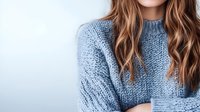Young Caucasian woman with long wavy brown hair wearing cozy blue knitted sweater against light background, fashion and lifestyle concept for winter season.