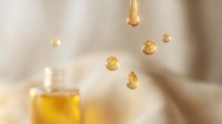 Golden oil droplets suspended in air above a bottle of amber liquid on soft neutral background, suitable for wellness, skincare, cosmetics and beauty product marketing.