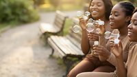 Happy African American women friends blowing soap bubbles together on park bench during sunny day outdoor leisure activity creating joyful memories.