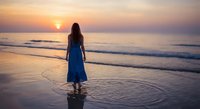 Woman in blue dress standing on beach watching golden sunset over ocean waves reflecting on wet sand creating peaceful moment.