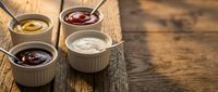 Four assorted dipping sauces in white ramekins on rustic wooden table, featuring ketchup, mustard, mayo, and BBQ sauce with spoons.