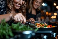 Two women with blonde and dark hair, smiling while cooking together at night, stirring colorful vegetables in a wok with bokeh lights in background. Suitable for cooking blogs, lifestyle content, and culinary websites.