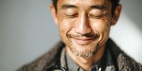 Close-up portrait of mature Asian man with eyes closed and gentle smile, expressing serenity and contentment against neutral background.