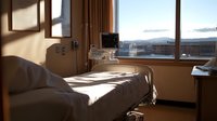 Empty hospital room with dramatic sunlight streaming through window, casting shadows across medical bed. City view and medical equipment visible in warm evening light.